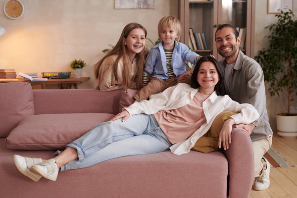 Family photo of a dark-haired mother with a young son on the sofa and a smiling husband and teenage daughter behind the sofa in a bright living room