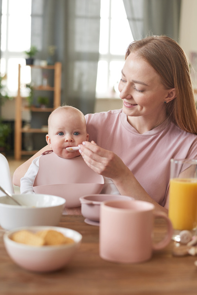 A young blonde mom feeds a baby in a pink bib with a pocket from a spoon sitting at the table