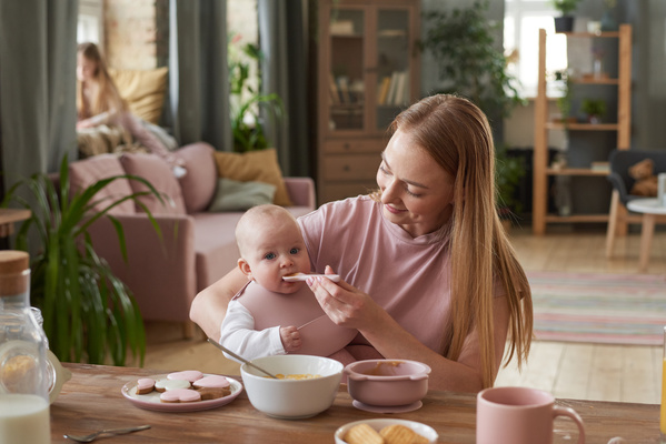 A woman with long blonde hair smiling spoon feeds her baby in a pink silicone bib with a pocket sitting at the table