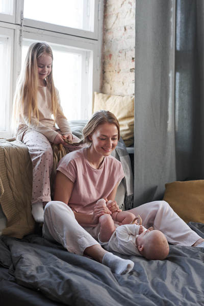 A girl with blonde long hair sitting on the windowsill braids her mothers hair while she entertains the baby