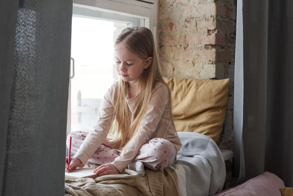 A girl with long blonde hair dressed in pajamas sitting on the windowsill with pillows and blankets draws with a red pencil