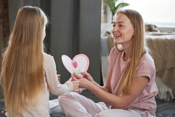 An excited mom sitting on the couch receives a heart-shaped card with a flower applique from her blonde daughter