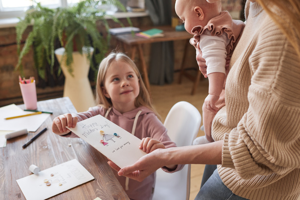 A daughter with blonde hair sitting at a table presents a mother with a baby in her arms with a Mothers day postcard with a drawing and an applique of buttons
