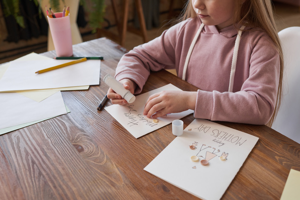 A girl glues buttons with glue stick on a homemade postcard with a picture for Mothers Day