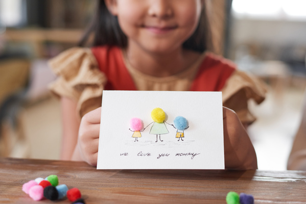 A girl holds a homemade postcard for Mothers Day with a pattern and applique of multicolored pompoms sitting at a table with materials for creativity