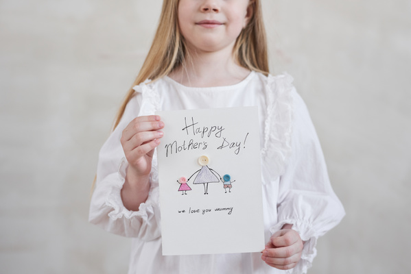 A card in honor of Mothers Day with an illustration and an applique of buttons is held by a girl with long blonde hair dressed in white clothes with ruffles