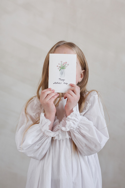 A blonde girl dressed in a white dress with ruffles holds a handmade postcard in honor of Mothers Day with a picture and congratulations covering her face with it