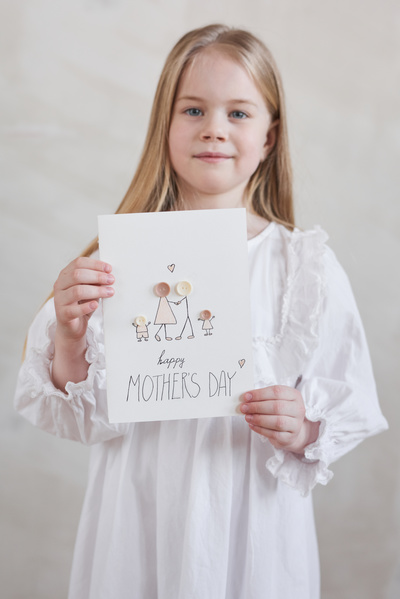 A girl with long blonde hair dressed in a white dress with ruffles shows a Mothers Day card with an illustration and an applique of buttons
