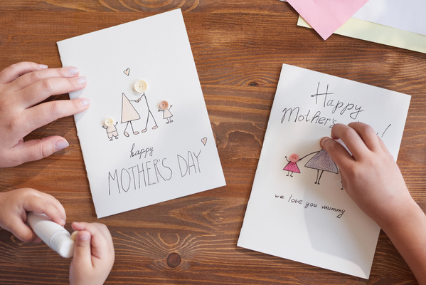 Close-up of homemade postcards for Mothers Day with illustrations on which mom and children glue button applications using glue stick