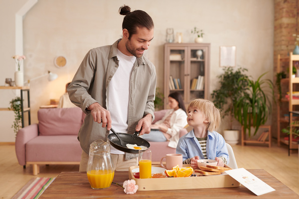 A man with his hair in a bun prepares a festive breakfast for mom looking at his son and laying the fried eggs on a plate on a tray