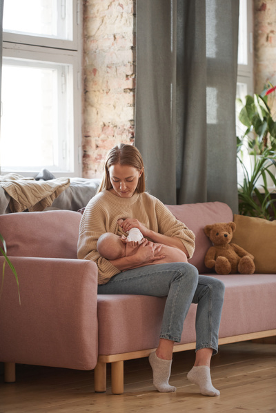 A blonde woman in a beige sweater and jeans sitting on the sofa in the living room breastfeeds a baby