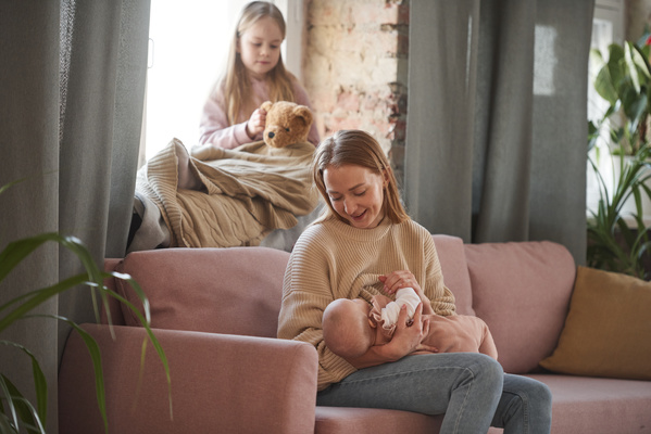 Blonde mother in light brown and jeans sitting on the sofa in the living room breastfeeding a kid
