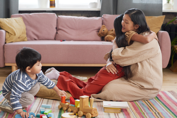 Mom hugs her dark-haired daughter tightly and the baby plays with wooden toys nearby