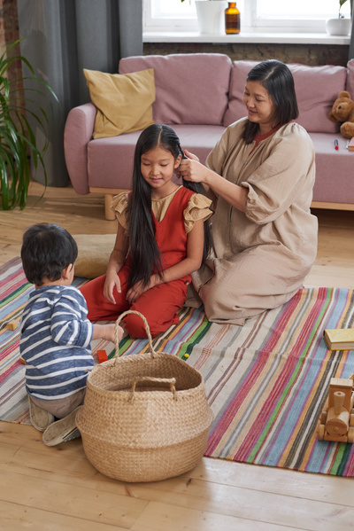 A woman braids her dark-haired daughters hair while sitting on the floor near a toddler playing with toys