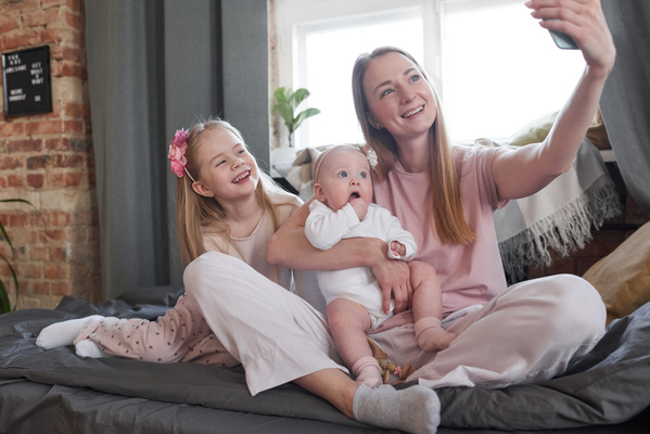A woman with blonde hair sitting with her daughter with a headband and a toddler on the sofa taking a selfie on her phone