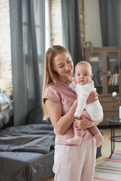 Mom with long blonde hair dressed in home clothes standing in the living room and holding a baby in a white bodysuit