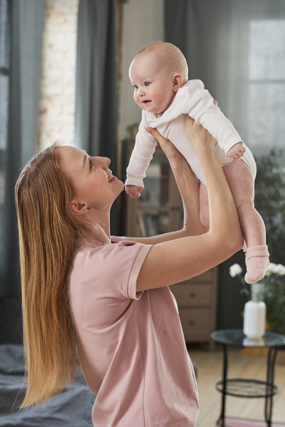 A mom with long blonde hair dressed in pajamas holds a baby in a white bodysuit on her outstretched arms