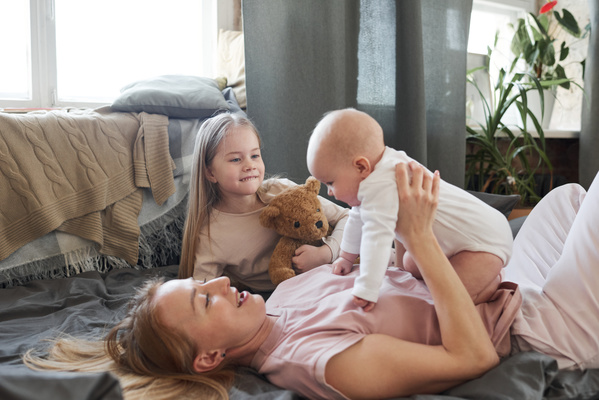 A woman plays with a baby sitting on her stomach lying on the couch with a blonde daughter in pajamas