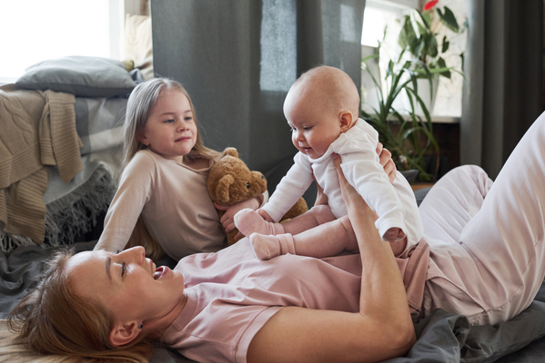 A girl with blonde hair and a teddy bear in her hands watches her mother playing with the baby lying next to her on the couch