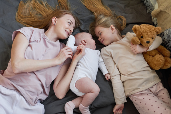A woman with long blonde hair lying on the sofa with a baby and an older daughter with a teddy bear in her hands dressed in pajamas