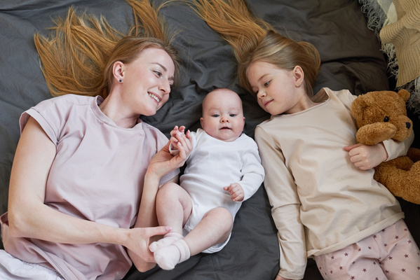 A mom with long blonde hair lying on the couch with a toddler and an older daughter with a teddy bear in her hands dressed in pajamas