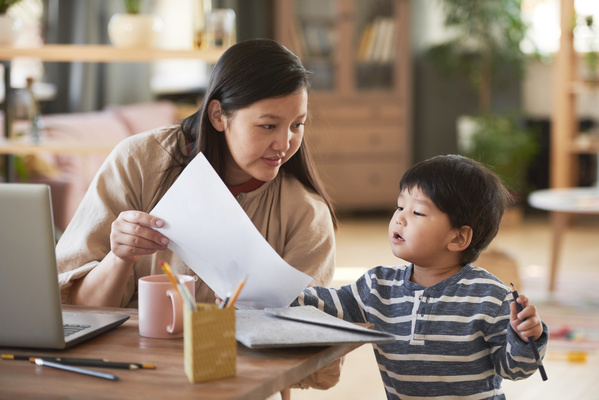 A dark-haired woman sitting at a table with a laptop gives a sheet of paper to little son with a pencil in hand