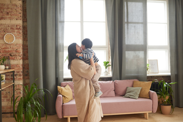 A woman in a beige tunic picking up her son in a striped jacket kisses him standing in a bright living room