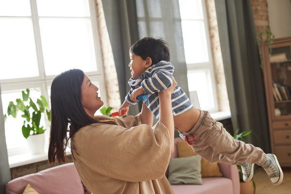 A woman in a beige tunic holding a laughing son in a striped T-shirt with long sleeves entertains him standing in a bright living room