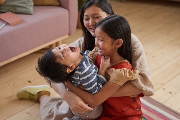 A dark-haired woman sitting on the floor smiling hugs laughing children
