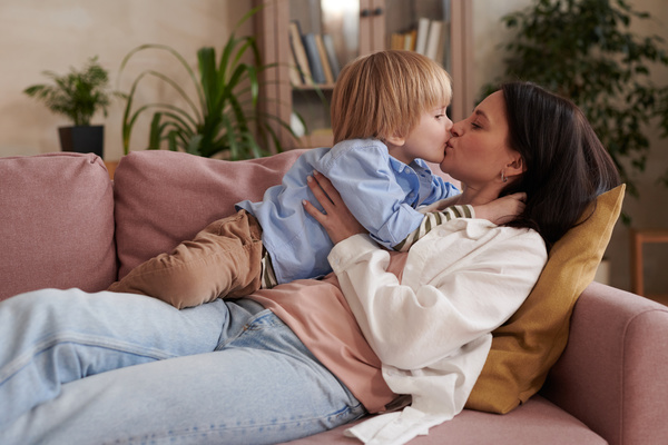 Dark-haired mom lying on a pink sofa in the living room kisses her blonde son