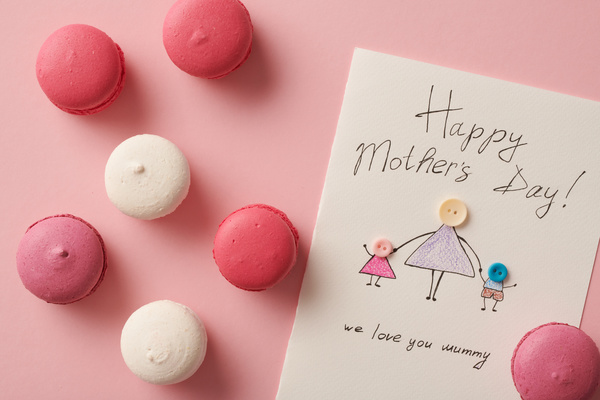 Pink and white macaroons and a handmade postcard for Mothers Day with an illustration and an applique of buttons are on the pink surface