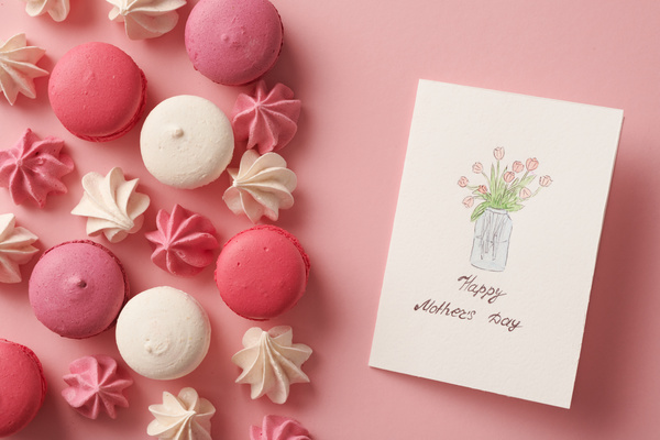 Pink and white macaroons and meringues and a homemade postcard in honor of the Mothers Holiday with an illustration are on the pink surface