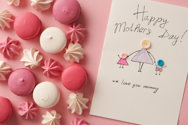 Top view of pink and white macaroons and meringues and a handmade Mothers Day card with an illustration on a pink surface