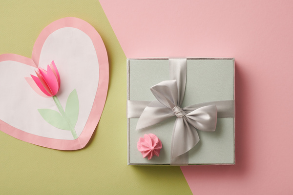 Handmade heart-shaped Mothers Day card and a gift box with a bow and meringue lying on a pink-green background