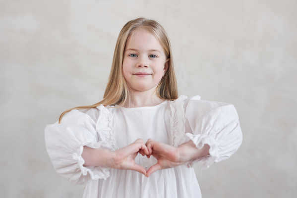 A girl with long blonde hair dressed in a white dress with ruffles shows a heart folded from her hands