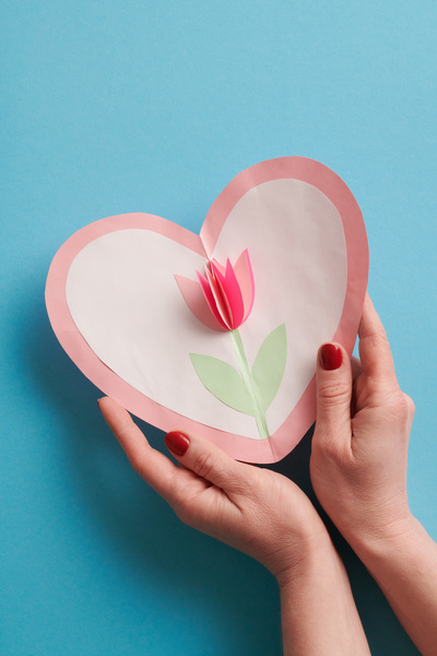 Homemade card in the shape of a heart decorated with a paper flower in female hands on a blue background