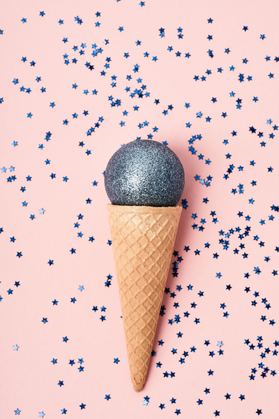 A shiny blue Christmas ball in a waffle cone resembling ice cream is located on a pink surface with a scattering of blue stars