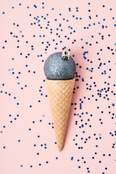 A blue glittered Christmas ball with a crown fastener in a waffle cone resembling ice cream is on a pink surface strewn with blue stars