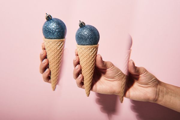 Futuristic image of a blue glittered Christmas ball with a crown fastener in a waffle cone resembling an ice cream held in hand on a pink background