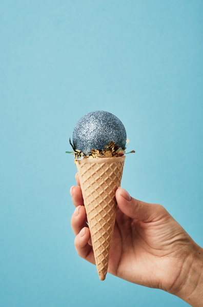 A blue shining Christmas ball is on a golden confetti in a waffle cone that is held on a blue background resembles ice cream