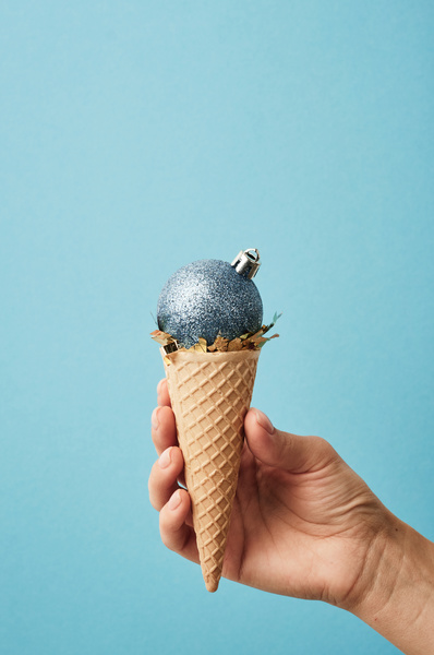 A blue shiny Christmas tree ball with a crown fastener is located on a golden confetti in a waffle cone and resembles ice cream that is held on a blue background