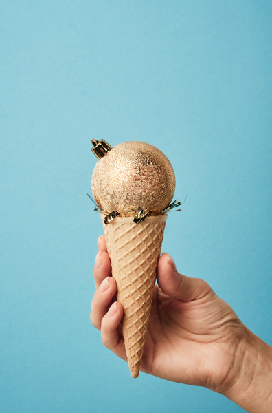 A golden frosted Christmas ball with crown fastener on a waffle cone filled with gold confetti resembling ice cream which is held on a blue background
