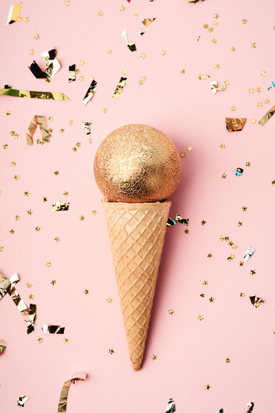 A frosted golden Christmas ball in a waffle cone that is in the hand on a pink background strewn with golden confetti and stars