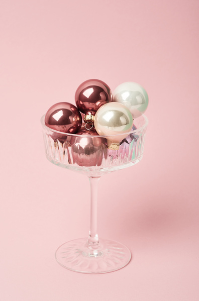 Pink and white glossy Christmas tree balls of small size lie in a champagne glass on a high leg on a pale pink background