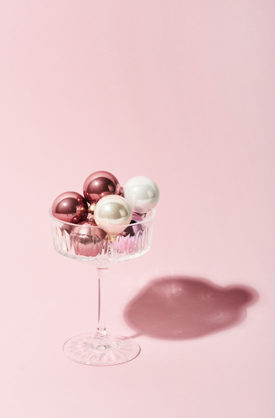 Pink and white glossy small Christmas balls in a champagne glass on a high leg which casts a shadow on a soft pink background