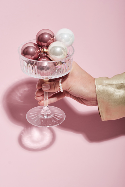 A champagne glass filled with pink and white Christmas balls in a female hand with a pearl ring on her thumb on a light pink background