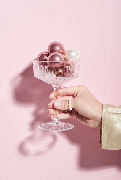 A high-stemmed glass filled with pink and white glossy Christmas balls in a womans hand with a pearl ring on her thumb on a light pink background