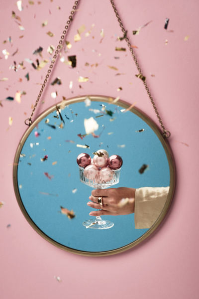 Confetti falls on a crystal glass filled with small pink and white Christmas balls in a female hand reflected in a round mirror hanging on a pink wall