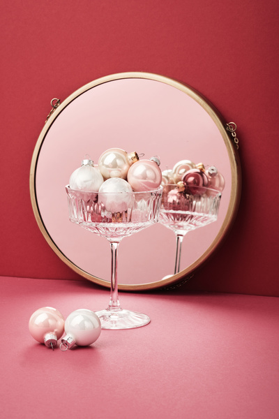 A wide glass with pink and white Christmas tree balls of small size standing on a pink surface next to two Christmas tree toys is reflected in a round mirror