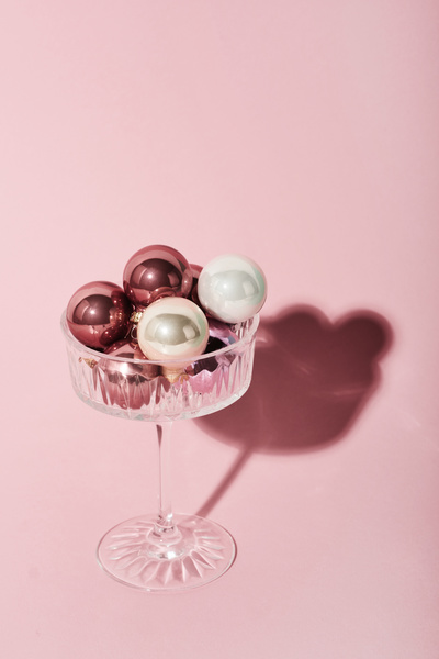 Small Christmas balls of white and pink with a glossy coating lie in a wide glass on a high stem which stands on a light pink surface casting a shadow